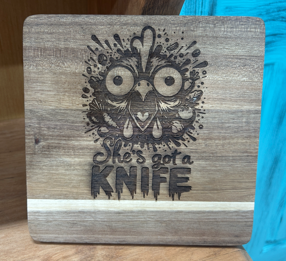 Square 8 x 8 inch acacia wood trivet with an engraved wide-eyed owl and the words She's Got a Knife