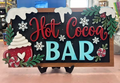 Hand-painted Hot Cocoa Bar sign with red and turquoise lettering, peppermint accents, greenery, and a cocoa mug.