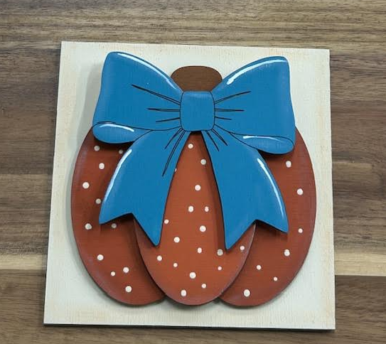 Hand-painted rust-orange pumpkin with cream dots and oversized blue bow on a square frame insert