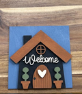 Finished Welcome House frame insert with a black house, brown roof and door, white heart and lettering, green topiaries, and blue background.