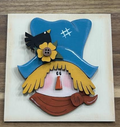 Hand-painted scarecrow frame insert with blue hat, yellow hair, rust-colored scarf, flower, and black bird
