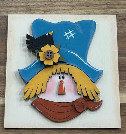 Hand-painted scarecrow frame insert with blue hat, yellow hair, rust-colored scarf, flower, and black bird