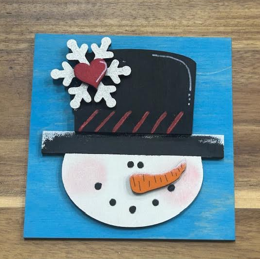 Finished snowman frame insert with a black top hat, white snowflake, red heart, and orange carrot nose on a blue background