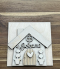 Unfinished Welcome House birch frame insert with a round window, heart-accented door, and two potted topiaries.