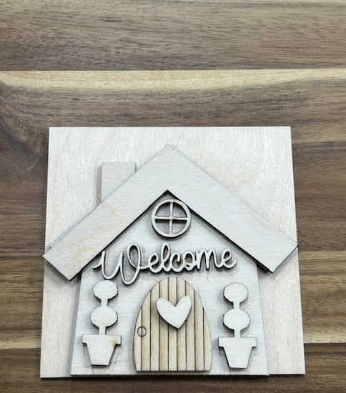 Unfinished Welcome House birch frame insert with a round window, heart-accented door, and two potted topiaries.